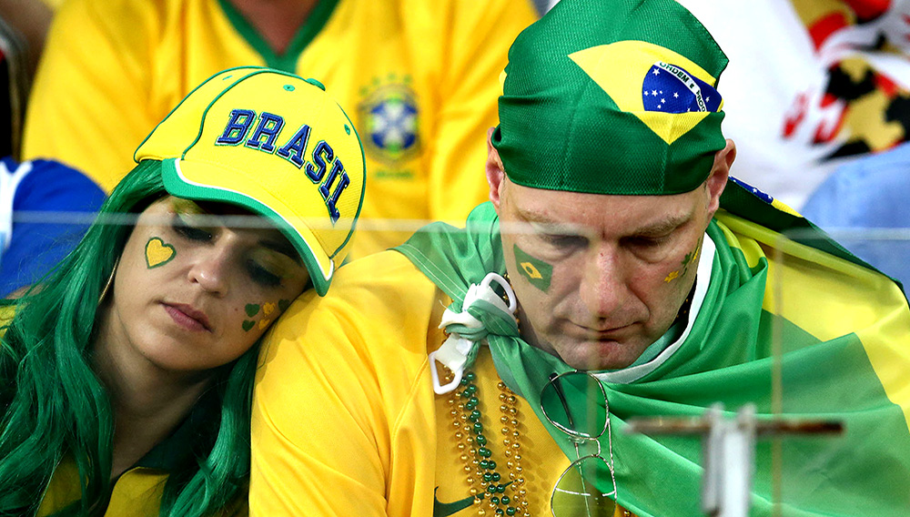 Dos hinchas adultos de la selección de Brasil, hombre y mujer en primer plano, vestidos con los colores verde y amarillo y con la mirada baja.