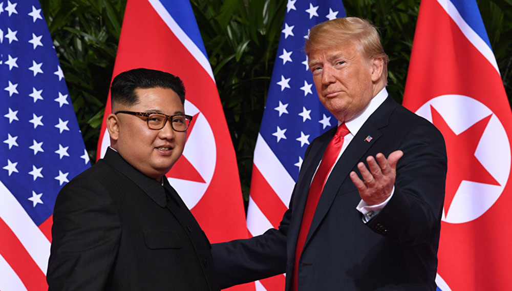 Delante de las banderas de Corea del Norte y Estados Unidos, Donald Trump hace un gesto con la mano izquierda invitando a un sonriente Kim Jong Un a mirar a las cámaras.