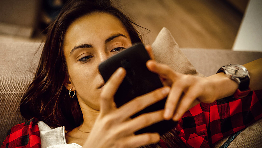 Mujer joven de cabellos largos y camisa roja y negra observa con atención su teléfono inteligente recostada en un mueble.