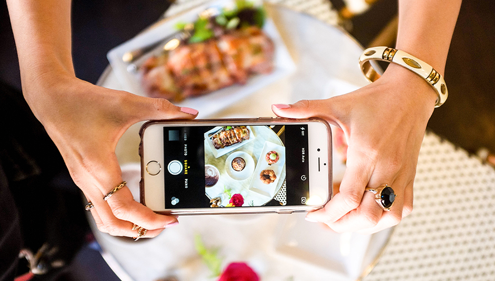 Manos de una mujer tomando una fotografía a un plato de comida con un smartphone desde arriba. Foto: cyneats