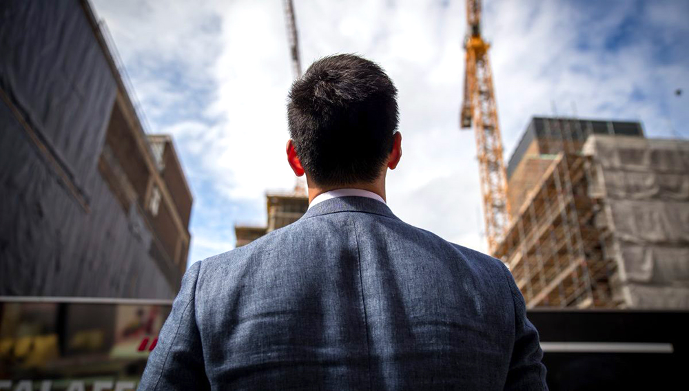 Hombre joven de espaldas a la cámara, mirando de lejos a unos edificios.