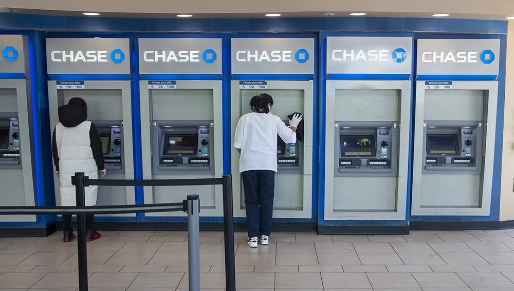 FILE - In this March 24, 2020 file photo, a Chase bank employee, right, disinfects the branches ATMs to fend off coronavirus as a customer uses another in the Flushing neighborhood of the Queens borough of New York. (AP Photo/Mary Altaffer, File)