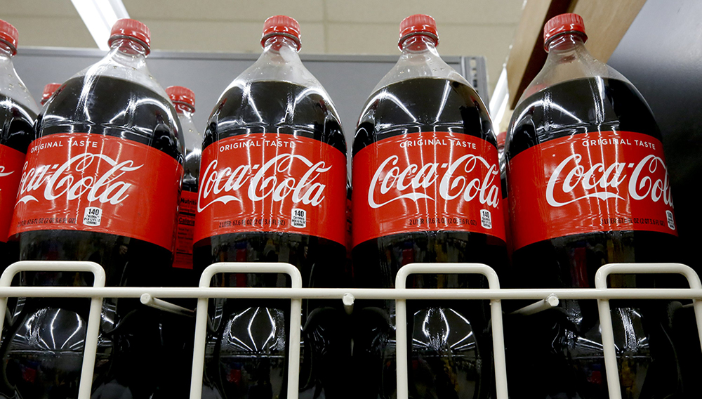 FILE- In this Aug. 8, 2018, file photo bottles of Coca-Cola sit on a shelf in a market in Pittsburgh. The Coca-Cola Co. said on Thursday, Oct. 22, 2020, it saw gradual improvement in the third quarter, as it turned its focus to emerging leaner from the global pandemic. Atlanta-based Coke said its revenue fell 9% to $8.7 billion. (AP Photo/Gene J. Puskar, File)