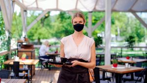 Portrait of waitress with face mask serving customers outdoors on terrace restaurant. | PHOTO: Halfpoint | Getty Images/iStockphoto
